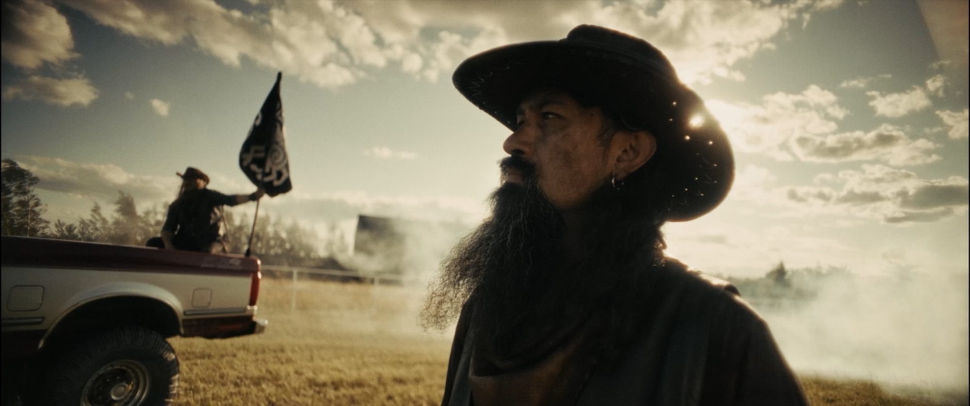 Profile of a long-bearded man in a cowboy hat; behind him a rider waves a black flag from a pickup at golden hour - Amon Amarth